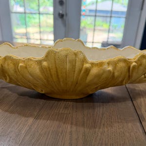 May include: An elongated, oval-shaped ceramic bowl featuring a textured, mustard-yellow exterior with a scalloped, shell-like design. The bowl has small, curved handles on each end and a smooth, cream-colored interior finish with a wavy rim.