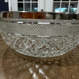 May include: A clear glass bowl featuring a detailed diamond-cut pattern across its exterior surface. The bowl is finished with a smooth, polished silver-tone metal rim that encircles the top edge, providing a contrast to the textured glass body.