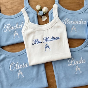 May include: A white tank top embroidered with Mrs. Madison and two champagne flutes in blue, surrounded by four light blue tank tops embroidered with the names Rachel, Alexandra, Olivia, and Linda, each featuring two champagne flutes.