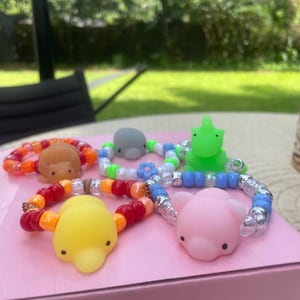 May include: A collection of colorful beaded bracelets featuring animal-shaped charms. The bracelets include a hedgehog, a dolphin, a frog, a duck, and a pig. The beads are in various colors, including orange, red, yellow, blue, and clear.