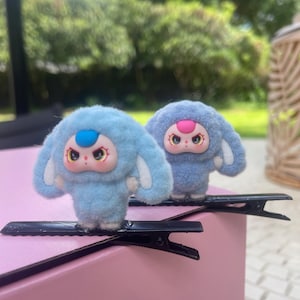 以下が含まれることがあります： ピンクの顔、大きな目、青またはピンクのトップスが特徴の漫画のキャラクターが付いた、2つのぬいぐるみブルーヘアクリップ。 クリップは黒いヘアクリップに取り付けられ、ぼやけた緑の背景のあるピンクの表面に配置されています。
