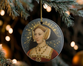 May include: A round ceramic ornament featuring a portrait of a person wearing a French hood and a red gown with gold trim. The figure is adorned with a multi-strand pearl necklace and a pendant. A golden starburst effect surrounds the head.