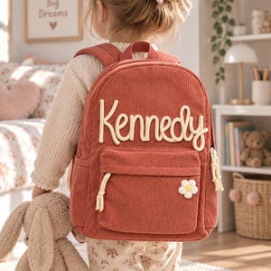 May include: A rust-colored corduroy backpack featuring the name Kennedy in raised cream-colored lettering across the top. The front pocket includes a cream-colored flower applique and braided rope zipper pulls on the main and front compartments.