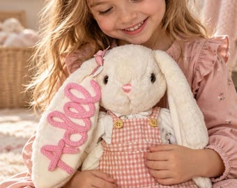 May include: A plush cream-coloured rabbit toy with long ears, wearing a pink and white gingham overall dress with two small buttons. The name Adlee is embroidered in pink script on one of the rabbit's ears, which also features a small pink bow.