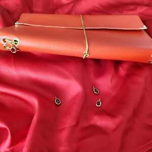 Può includere: Un diario in pelle arancione con chiusura a portafoglio, dotato di occhielli in metallo dorato e un ciondolo a forma di goccia rossa. Tre ulteriori ciondoli a forma di goccia rossa sono posizionati sciolti nelle vicinanze.