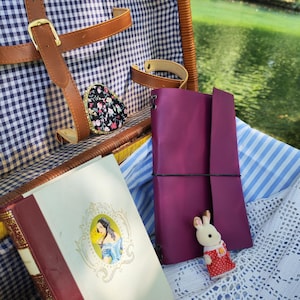 Puede incluir: Una cesta de picnic de mimbre con forro de cuadros vichy azul y blanco, que contiene un libro con un retrato en la portada, un diario de cuero morado sujeto con una banda elástica negra y una pequeña figura de conejo con vestido rojo a lunares.