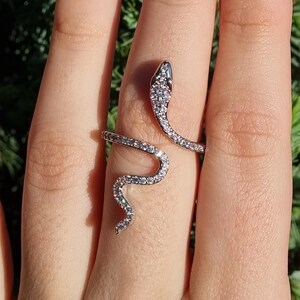 May include: A silver-toned wrap ring shaped like a snake, featuring a coiled body and head encrusted with numerous small, clear, sparkling stones. The metal band winds around the finger, with the snake's head and tail resting at different points.
