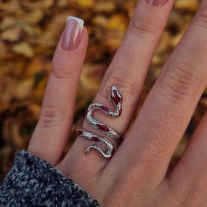 May include: A silver-toned snake ring featuring a textured metallic body and inlaid red enamel accents along the serpent's back. The ring wraps around the finger in a coiled design, showcasing a detailed head and tail with a polished finish.