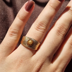 Könnte beinhalten: Ein breiter Ring aus goldfarbenem Metall mit einem zentralen, runden, bräunlich-goldenen Edelstein. Das Band ist mit einem strahlenförmigen Muster versehen, das in die Metalloberfläche eingraviert ist und vom Stein ausgeht.
