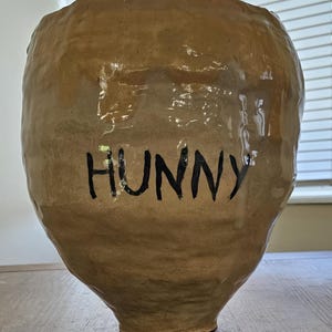 以下が含まれることがあります： 手作りの、薄茶色の陶器の壺で、黒い文字で「HUNNY」と書かれています。壺は広い開口部と丸みを帯びた胴体があり、小さな底に向かって先細りになっています。表面は質感のある素朴な外観をしています。