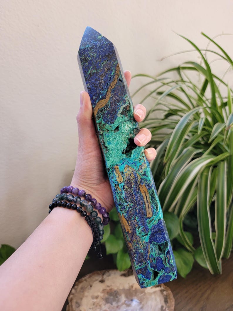 May include: A tall, polished azurite and malachite crystal tower featuring a vibrant mix of deep blue, bright green, and earthy brown mineral veins. The stone has a faceted, pointed top and a textured, natural surface with visible crystalline cavities.