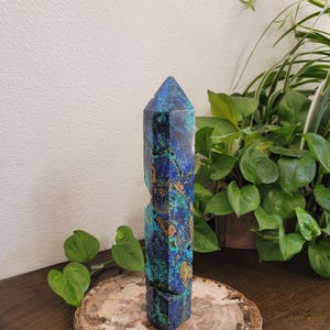 May include: A tall, faceted crystal tower featuring a vibrant mix of deep royal blue, turquoise, and earthy brown mineral inclusions. The stone has a polished, pointed top and a natural, rugged texture along its vertical sides, standing upright.