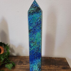 May include: A tall, rectangular crystal obelisk with a pointed top. The stone features a blend of blue and green hues, with darker veins running through the surface. The obelisk is standing on a wooden surface.