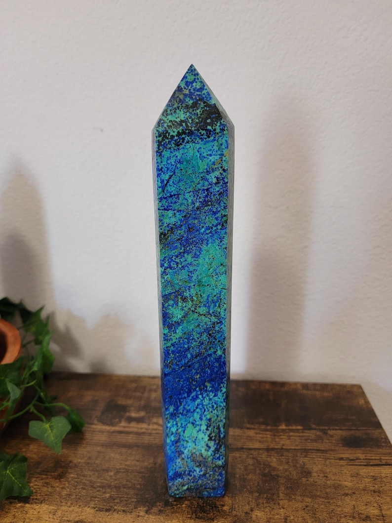 May include: A tall, rectangular crystal obelisk with a pointed top. The stone features a blend of blue and green hues, with darker veins running through the surface. The obelisk is standing on a wooden surface.