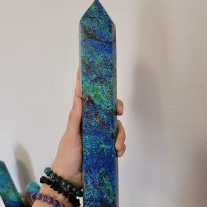 May include: A tall, rectangular crystal obelisk with a pointed top. The stone features a vibrant mix of blue, green, and brown hues. The obelisk is held in a hand, showcasing its size and texture. The crystal is a decorative item.