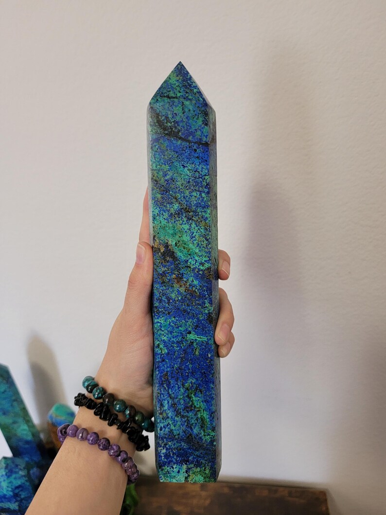 May include: A tall, rectangular crystal obelisk with a pointed top. The stone features a vibrant mix of blue, green, and brown hues. The obelisk is held in a hand, showcasing its size and texture. The crystal is a decorative item.