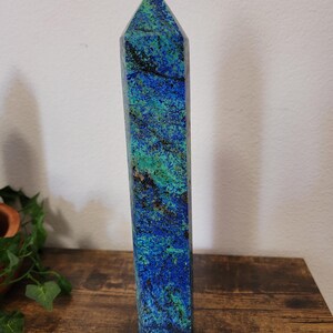 May include: A tall, rectangular azurite crystal obelisk with a pointed top. The stone features a vibrant mix of blue and green hues with dark veining. It stands upright on a wooden surface, showcasing its natural beauty and unique patterns.