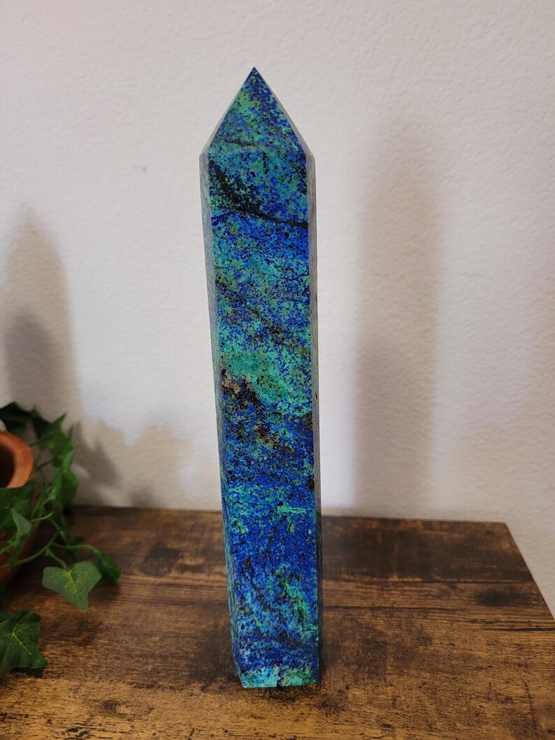 May include: A tall, rectangular azurite crystal obelisk with a pointed top. The stone features a vibrant mix of blue and green hues with dark veining. It stands upright on a wooden surface, showcasing its natural beauty and unique patterns.