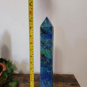 May include: A tall, rectangular azurite crystal obelisk, approximately 13 inches tall, with a pointed top. The stone features a mix of deep blue and green hues. A yellow measuring tape is placed next to the obelisk.