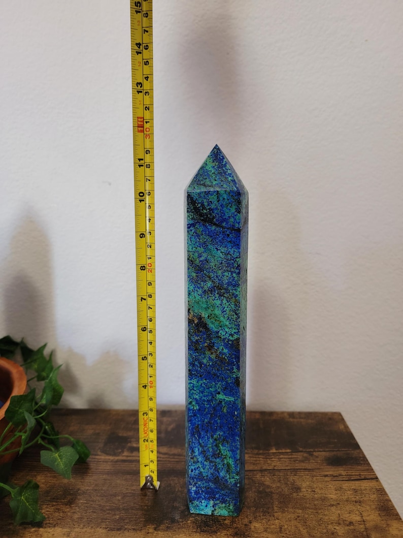 May include: A tall, rectangular azurite crystal obelisk, approximately 13 inches tall, with a pointed top. The stone features a mix of deep blue and green hues. A yellow measuring tape is placed next to the obelisk.