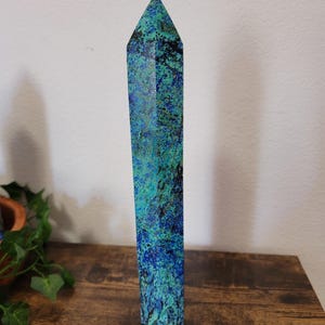 May include: A tall, rectangular crystal obelisk with a pointed top. The stone features a mottled pattern of deep blue, teal, and black. The obelisk is standing upright on a wooden surface.