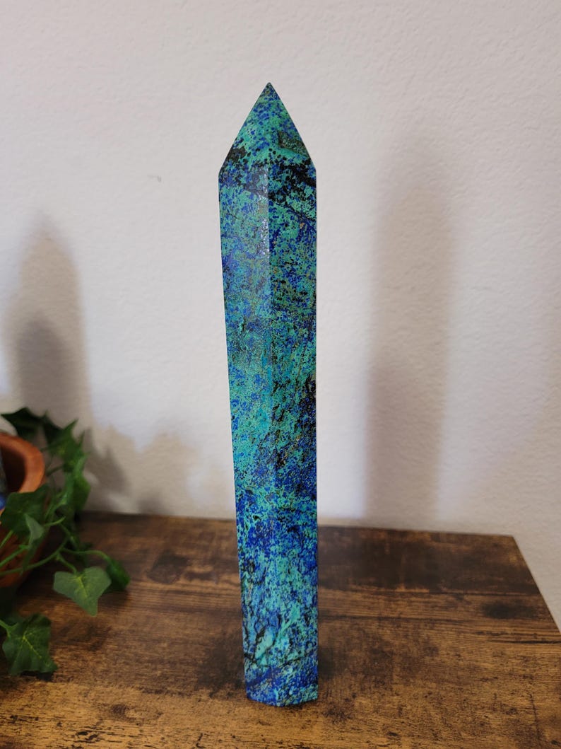 May include: A tall, rectangular crystal obelisk with a pointed top. The stone features a mottled pattern of deep blue, teal, and black. The obelisk is standing upright on a wooden surface.