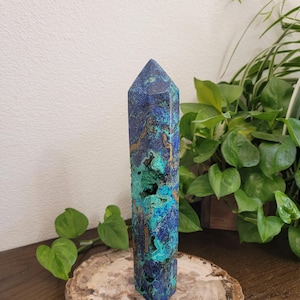 May include: A polished azurite and malachite crystal tower featuring a hexagonal point. The stone displays a vibrant mix of deep royal blue and bright turquoise green colors with natural brown matrix inclusions and small textured cavities.