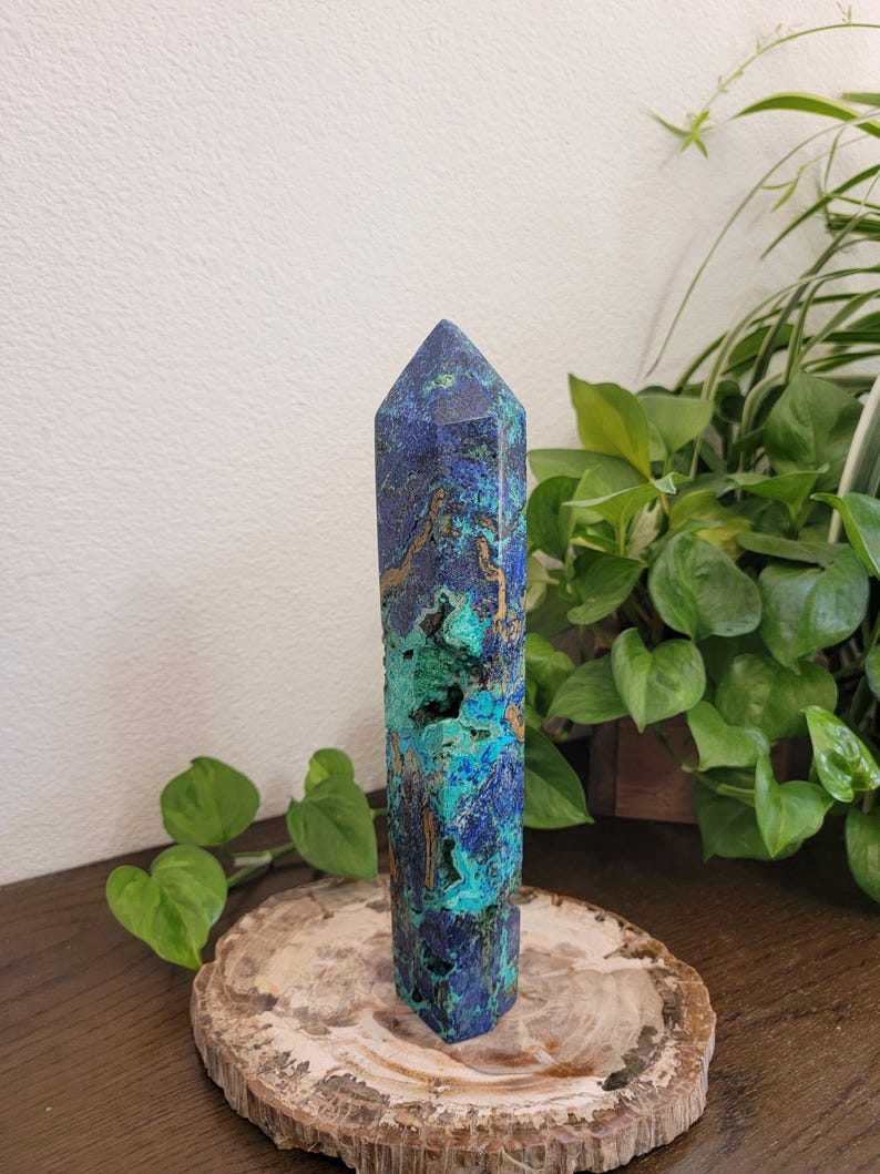 May include: A polished azurite and malachite crystal tower featuring a hexagonal point. The stone displays a vibrant mix of deep royal blue and bright turquoise green colors with natural brown matrix inclusions and small textured cavities.