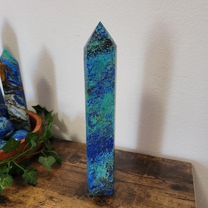 May include: A tall, rectangular azurite crystal point with a pointed top. The crystal features a mix of deep blue and teal colors with dark green and black accents. It stands upright on a wooden surface, with other crystals and greenery in the background.