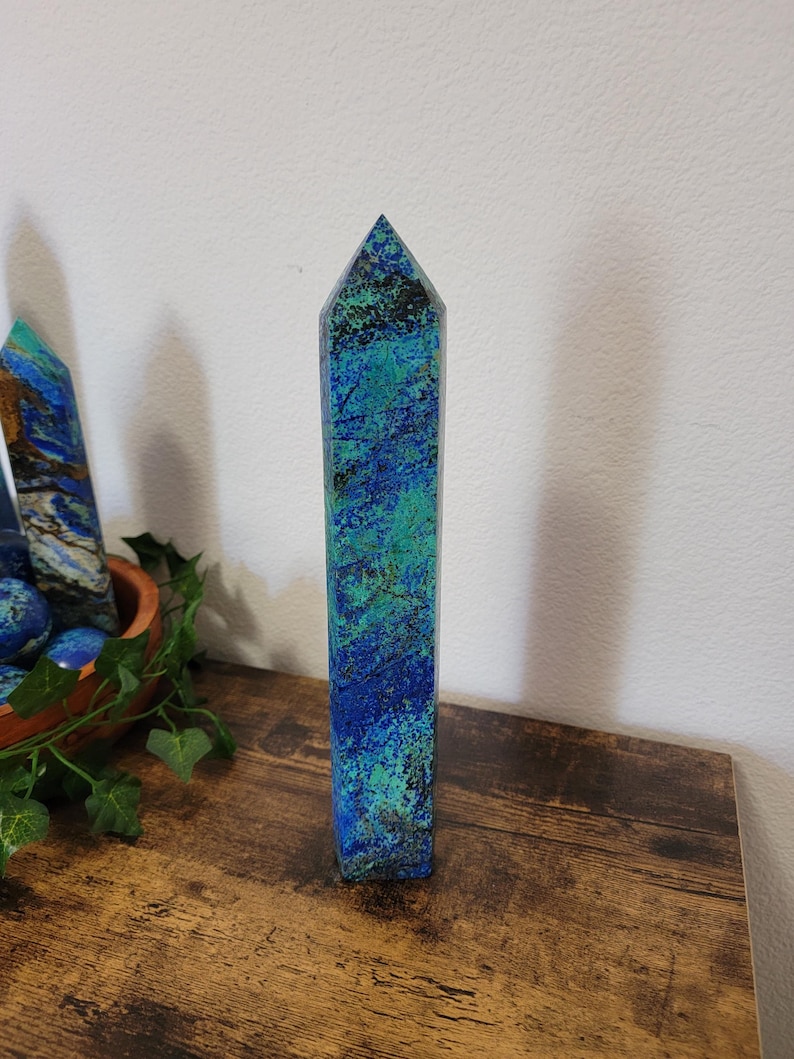 May include: A tall, rectangular azurite crystal point with a pointed top. The crystal features a mix of deep blue and teal colors with dark green and black accents. It stands upright on a wooden surface, with other crystals and greenery in the background.