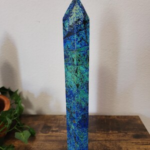 May include: A tall, rectangular azurite crystal obelisk with a pointed top. The stone features a mix of deep blue and teal green hues with dark veining. The obelisk is standing on a wooden surface.
