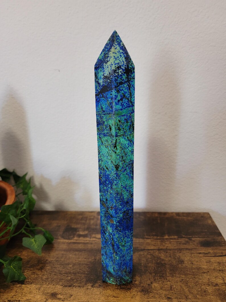 May include: A tall, rectangular azurite crystal obelisk with a pointed top. The stone features a mix of deep blue and teal green hues with dark veining. The obelisk is standing on a wooden surface.