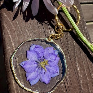 May include: A circular keychain featuring a pressed purple delphinium flower encased in clear resin. The pendant has a metallic gold-toned irregular edge and is attached to a gold-toned metal split ring by a short chain link connector.
