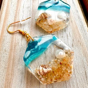 May include: A pair of hexagonal resin earrings featuring a beach scene with embedded sand, small seashells, and white pearls. The design includes a gradient of translucent blue and white waves, finished with gold-toned metal fishhook ear wires.