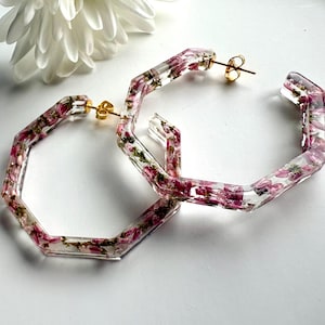 May include: A pair of clear, hexagonal hoop earrings featuring embedded pink and purple dried flowers. The earrings are constructed from a transparent resin material and include gold-toned metal post backings for secure attachment to the ear.