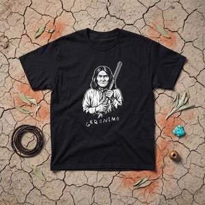 May include: A black short-sleeve t-shirt featuring a white graphic illustration of Geronimo holding a rifle. The text GERONIMO is printed in white below the figure. The shirt is displayed flat against a dry, cracked earth background.