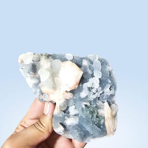 May include: A raw mineral specimen featuring a dark grey, translucent base with clusters of pale peach and white crystalline formations. The stone has a rough, textured surface with varied crystal shapes embedded throughout the dark grey matrix.