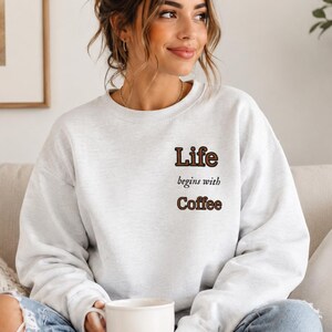 May include: A light gray sweatshirt with the text "Life begins with Coffee" in brown lettering. The sweatshirt is a crew neck style and is being worn by a person. The person is holding a white mug.