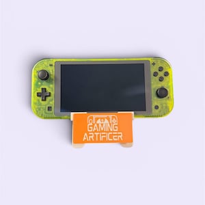 Puede incluir: Una consola de juegos portátil de color amarillo neón translúcido con botones y joysticks negros, apoyada sobre un soporte naranja con el texto blanco GAMING ARTIFICER y un pequeño icono de mando sobre la palabra gaming.