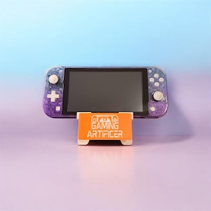 Op de afbeelding: Een paars en blauw verloop handheld spelcomputer met witte knoppen rust op een oranje standaard. De standaard is voorzien van een witte afbeelding van een controller en de tekst GAMING ARTIFICER in een vet, schreefloos lettertype op de voorzijde.