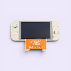 May include: A handheld gaming console with a light beige casing and a black screen, resting on an orange plastic stand. The stand features a white graphic logo with a stylized dragon icon and the text GAMING ARTIFICER printed in white.