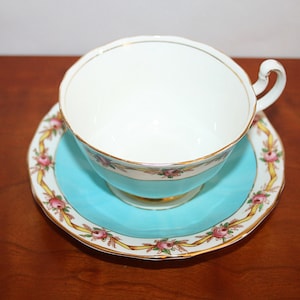 Puede incluir: Un juego de taza y platillo de té vintage con una base azul turquesa y un interior blanco. El platillo y la taza están decorados con un borde dorado y un patrón floral de rosas rosas y cintas amarillas.