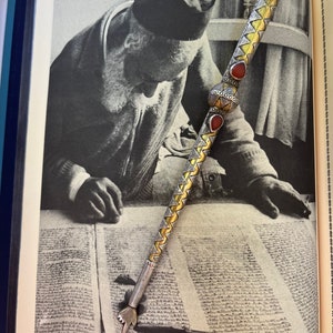 Może przedstawiać: Srebrny i złoty jad, żydowski wskaźnik religijny, z czerwonymi akcentami z kamieni szlachetnych. Jad leży na stronie książki z tekstem hebrajskim.