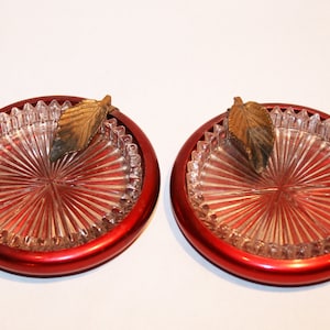 May include: Two red glass and metal coasters with a clear glass center and a gold leaf design.