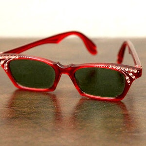 Op de afbeelding: Rode cat-eye zonnebril met strass op de monturen en donkergroene glazen.