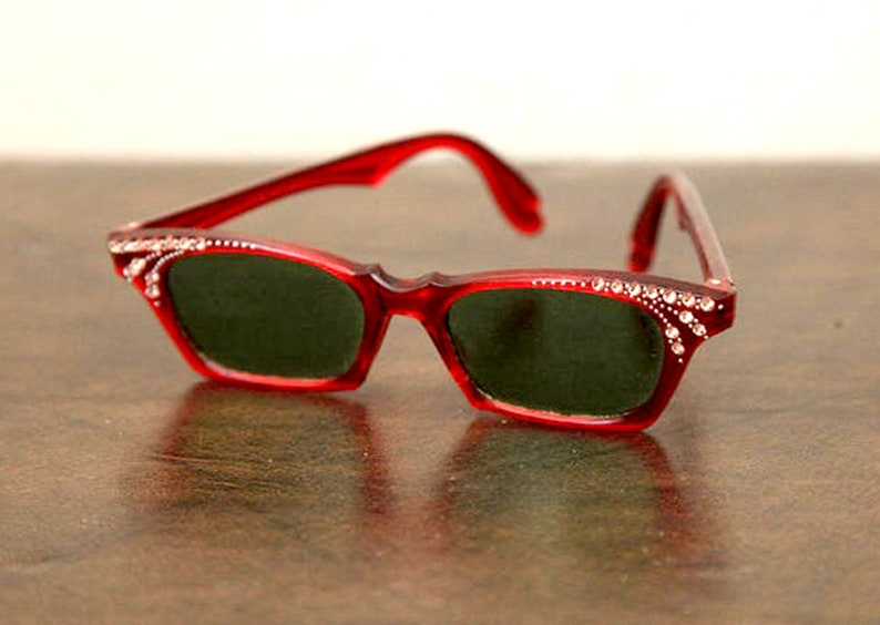 Op de afbeelding: Rode cat-eye zonnebril met strass op de monturen en donkergroene glazen.