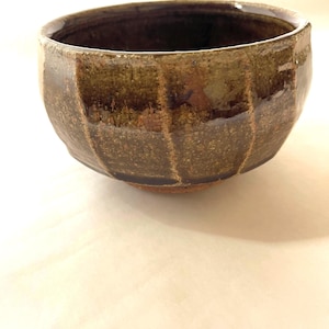May include: A round ceramic bowl with a dark brown and olive green mottled glaze. The exterior features vertical indentations creating a faceted texture, while the interior is smooth and dark. The base is unglazed, revealing a reddish-brown clay.