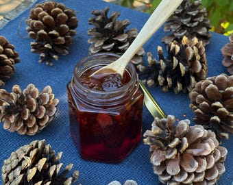 May include: A hexagonal glass jar filled with dark red jam containing visible fruit pieces, accompanied by a light-coloured wooden spoon resting inside. The jar is surrounded by several brown pine cones arranged on a textured blue fabric surface.