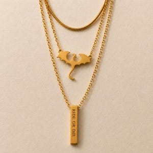 Pode incluir: Um colar em camadas de tom dourado com três correntes distintas. A corrente superior é uma corrente de cobra, a corrente do meio tem um pingente de silhueta de dragão e a corrente inferior apresenta um pingente de barra retangular vertical gravado com RIDE OR DIE.