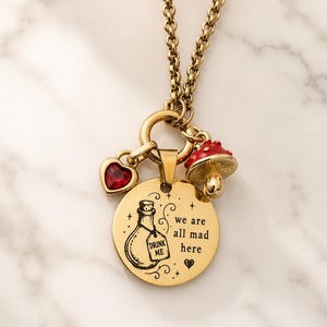 May include: A gold-toned necklace featuring a circular pendant engraved with a bottle illustration, the text "DRINK ME" and "we are all mad here," accompanied by a small red heart-shaped charm and a red and gold mushroom charm.
